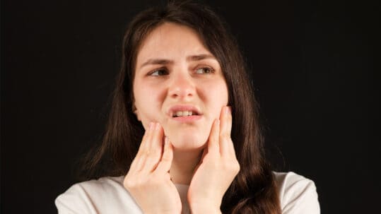 A young woman grimaces in pain and holds her jaw with her hands, implying TMJ pain, as she looks off to the side.