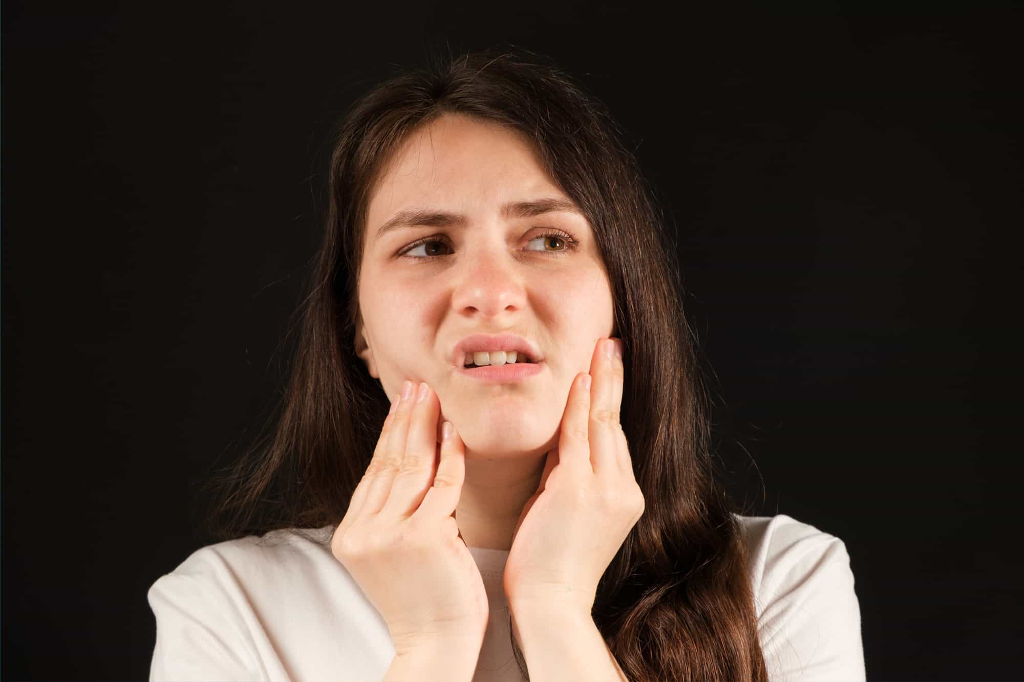 A young woman grimaces in pain and holds her jaw with her hands, implying TMJ pain, as she looks off to the side.