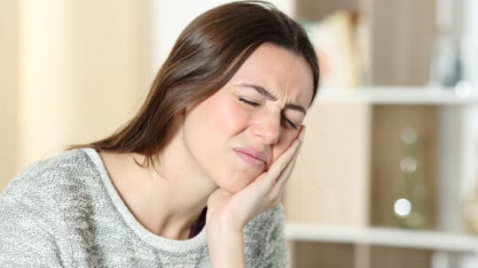 A young woman with brown hair and a sweater holds her jaw because of her TMJ pain.