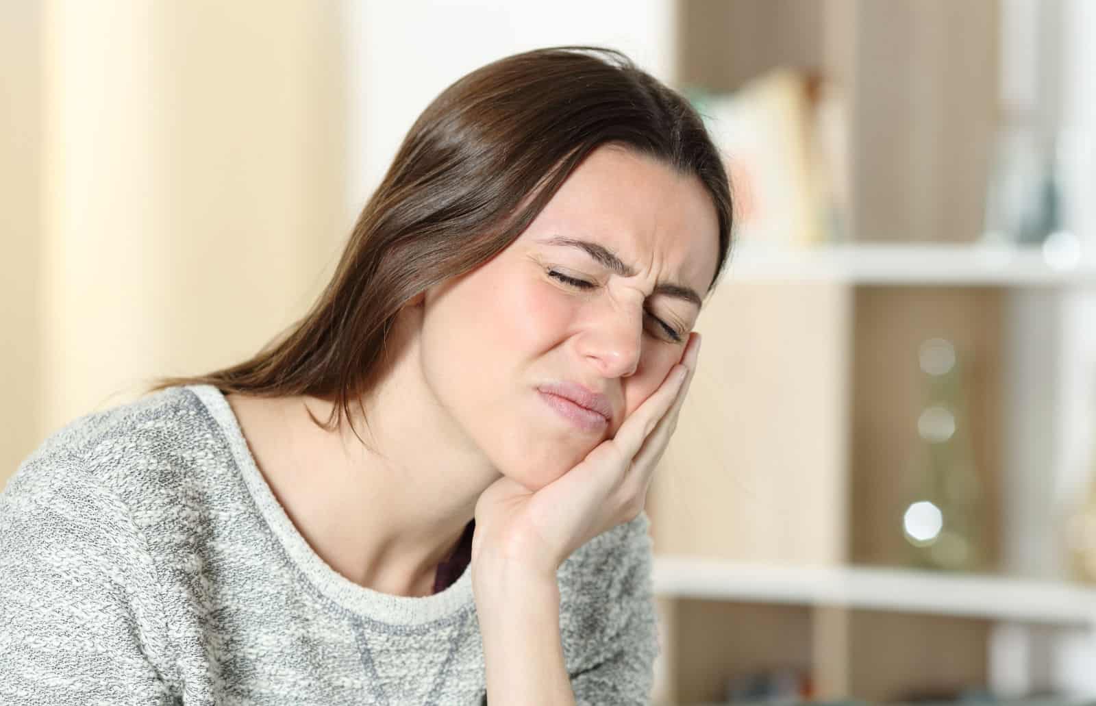 A young woman with brown hair and a sweater holds her jaw because of her TMJ pain.