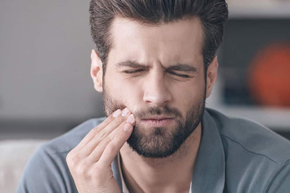 A man with brown hair and a gray shirt touches his face because of his TMJ pain.