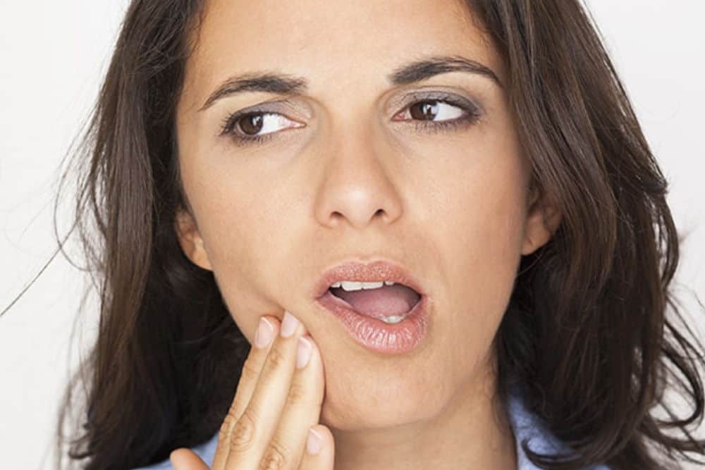 A woman with dark hair touches her jaw, which appears to be out of place and causing her TMJ pain.