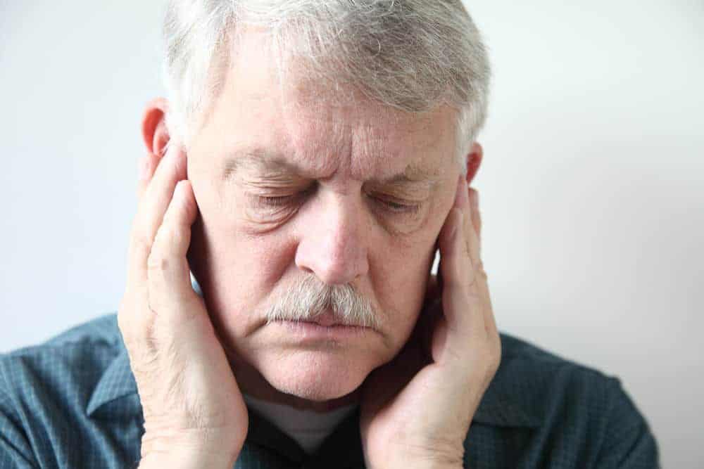 An older man in a blue shirt holds his hands over his jaw as he looks down, likely due to his TMJ pain.