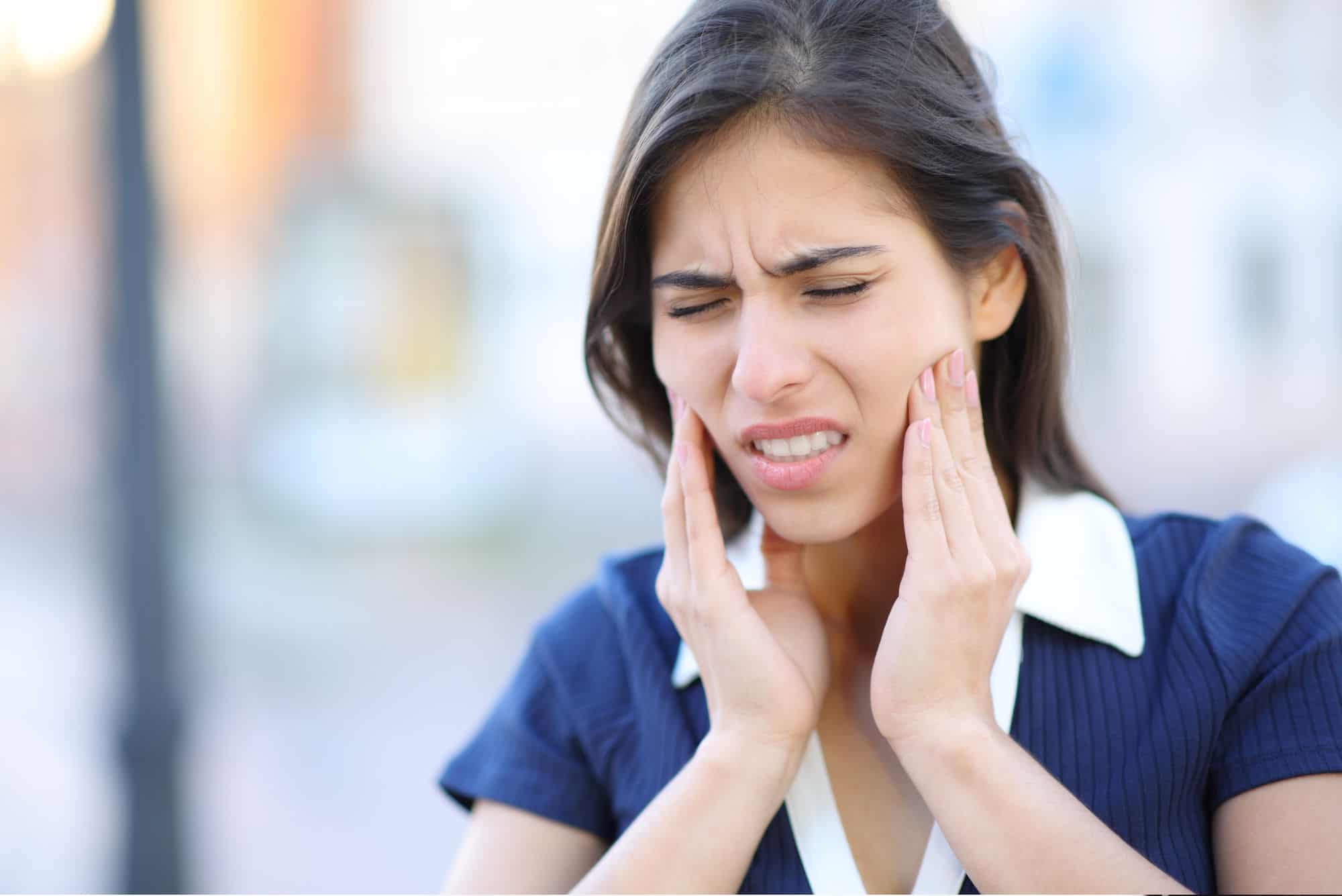 A pretty young woman grimaces and holds her jaw because of her TMJ pain. She's wearing a dark blue shirt with a white collar in front of a blurred background.