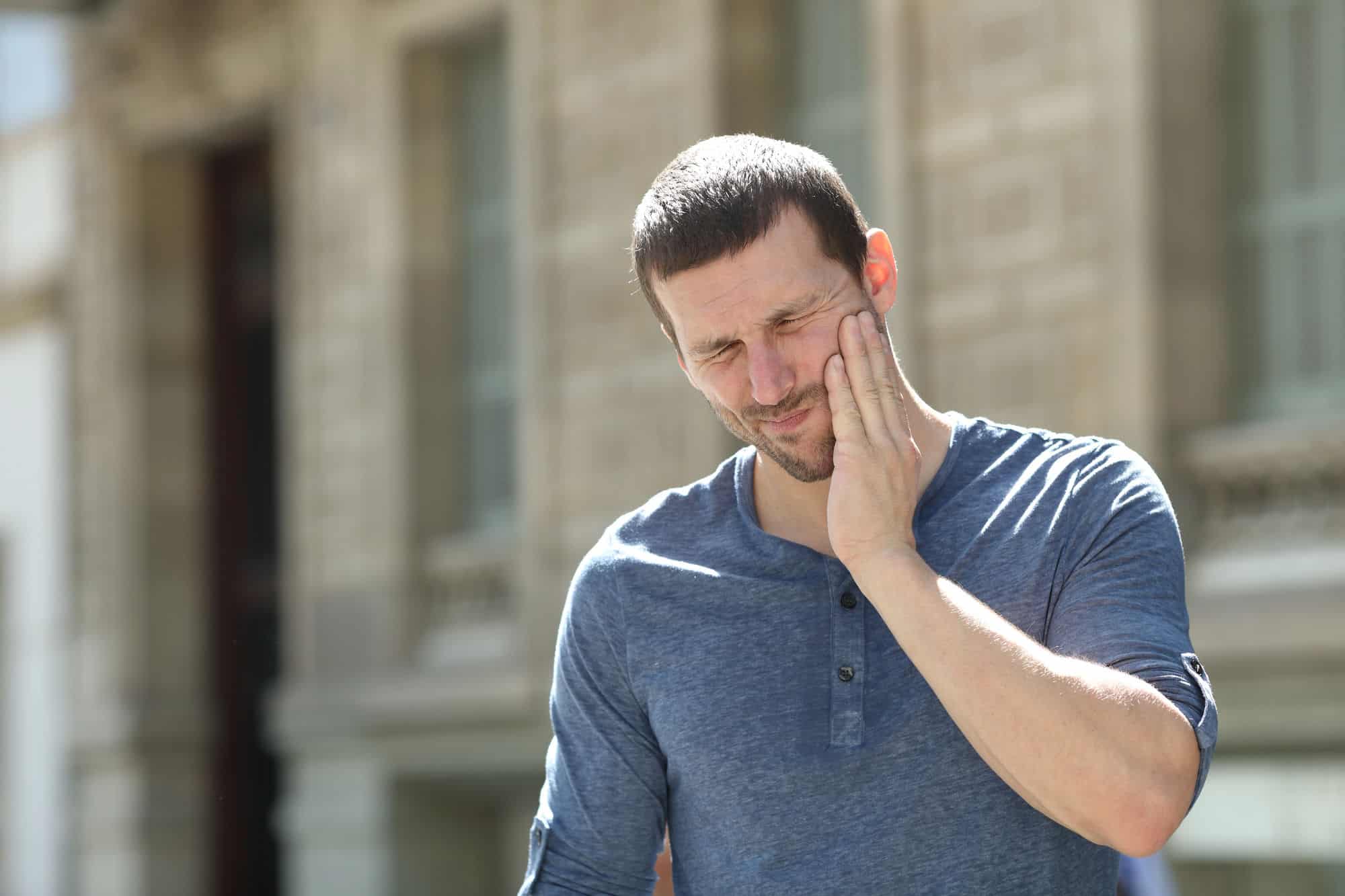 A man in a light blue Henley shirt holds his jaw because of his persistent TMJ pain in front of a blurred background.