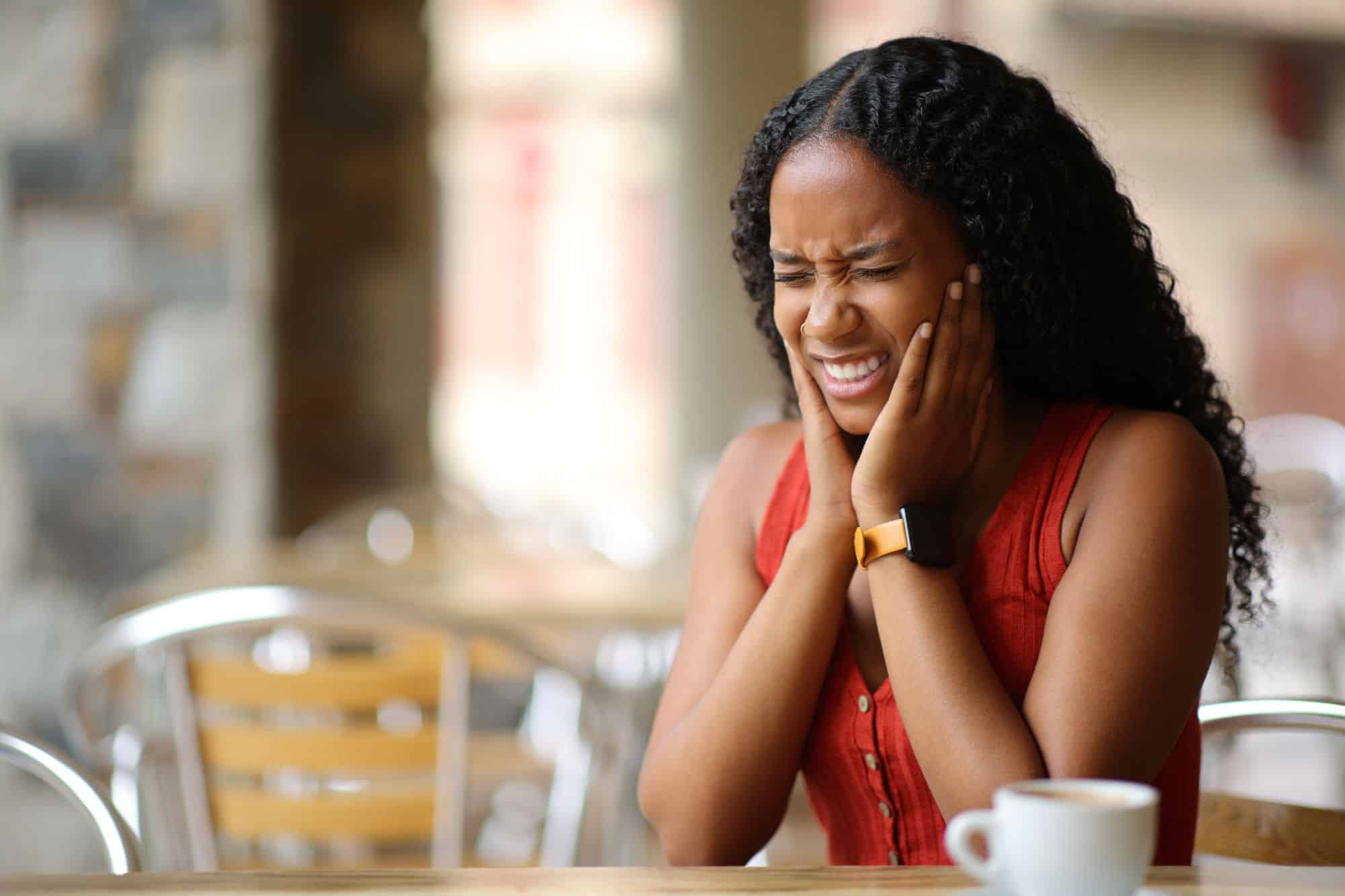 A young black woman holds her jaw because of her TMJ pain in a cafe with a blurred background.