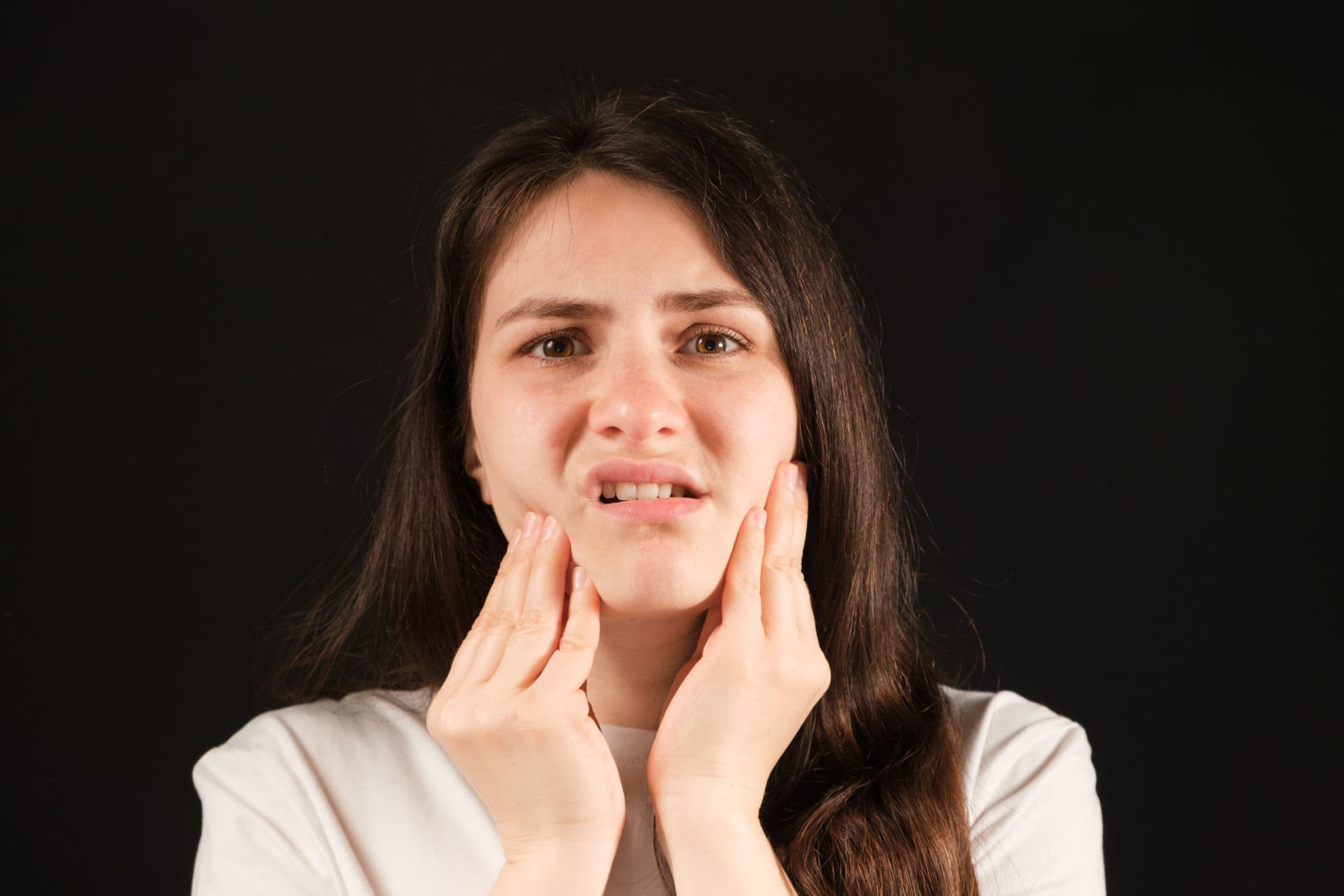 A young woman with TMJ pain holds her jaw in front of a black background.