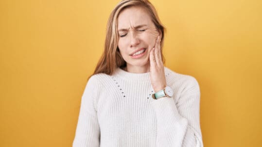 A young woman in a white sweater holds her aching jaw in front of a yellow background.