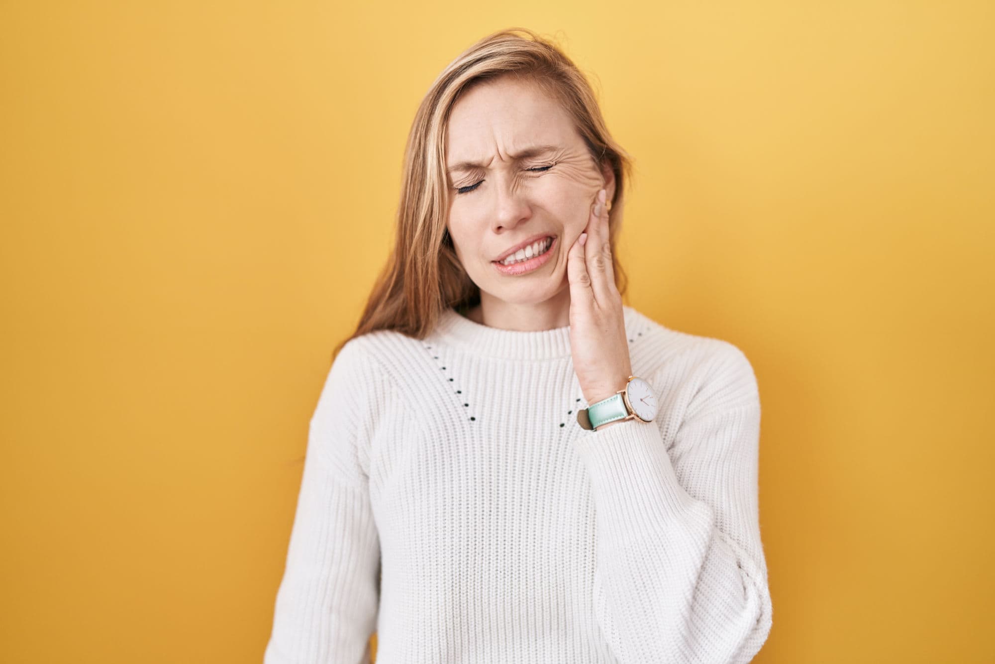 A young woman in a white sweater holds her aching jaw in front of a yellow background.