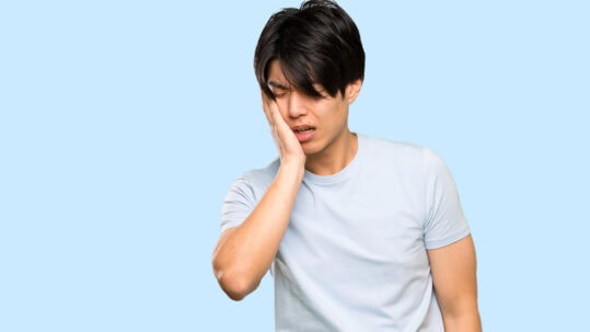 An Asian man in a blue shirt holds his jaw in pain in front of a blue background.