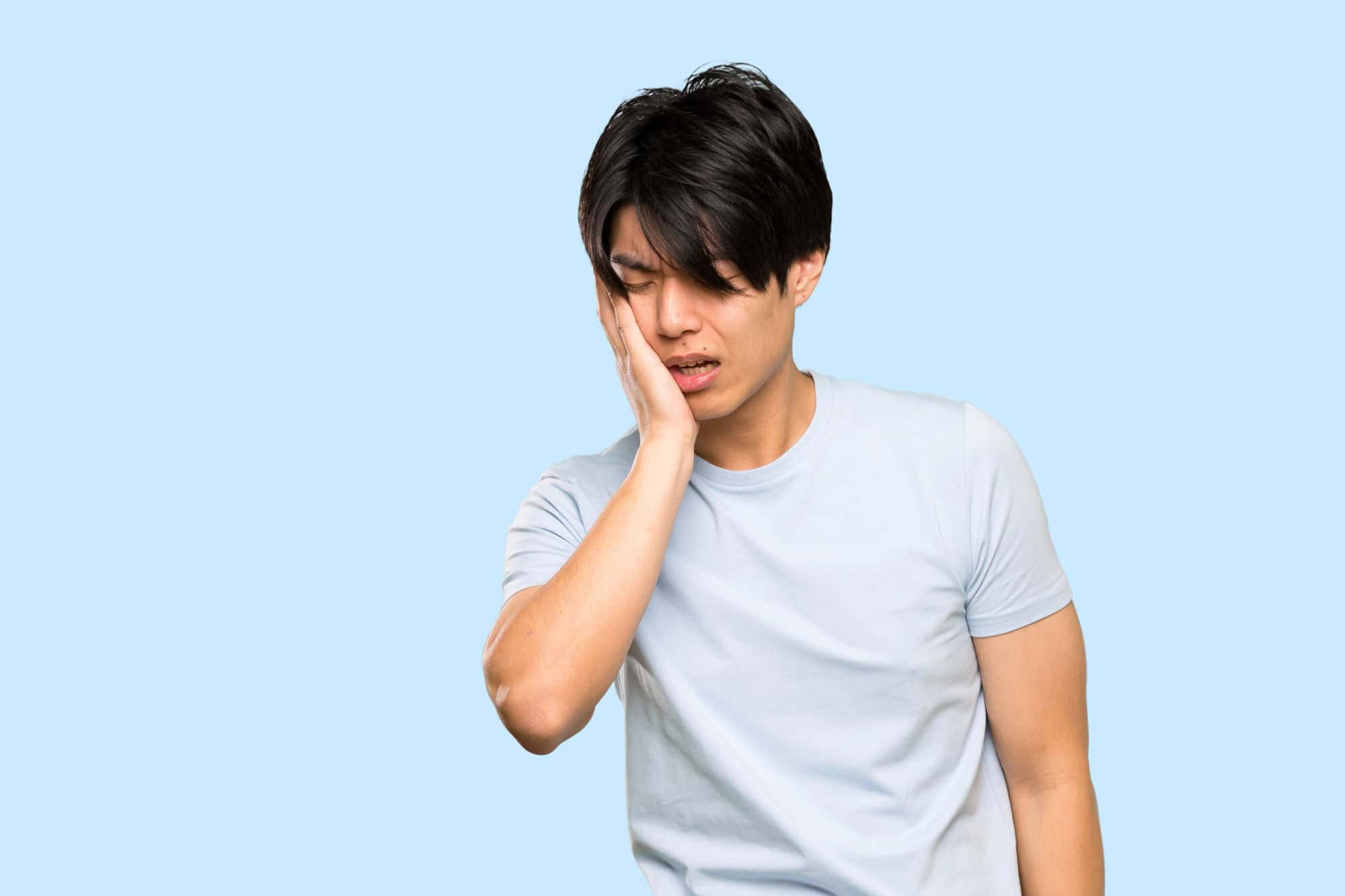 An Asian man in a blue shirt holds his jaw in pain in front of a blue background.