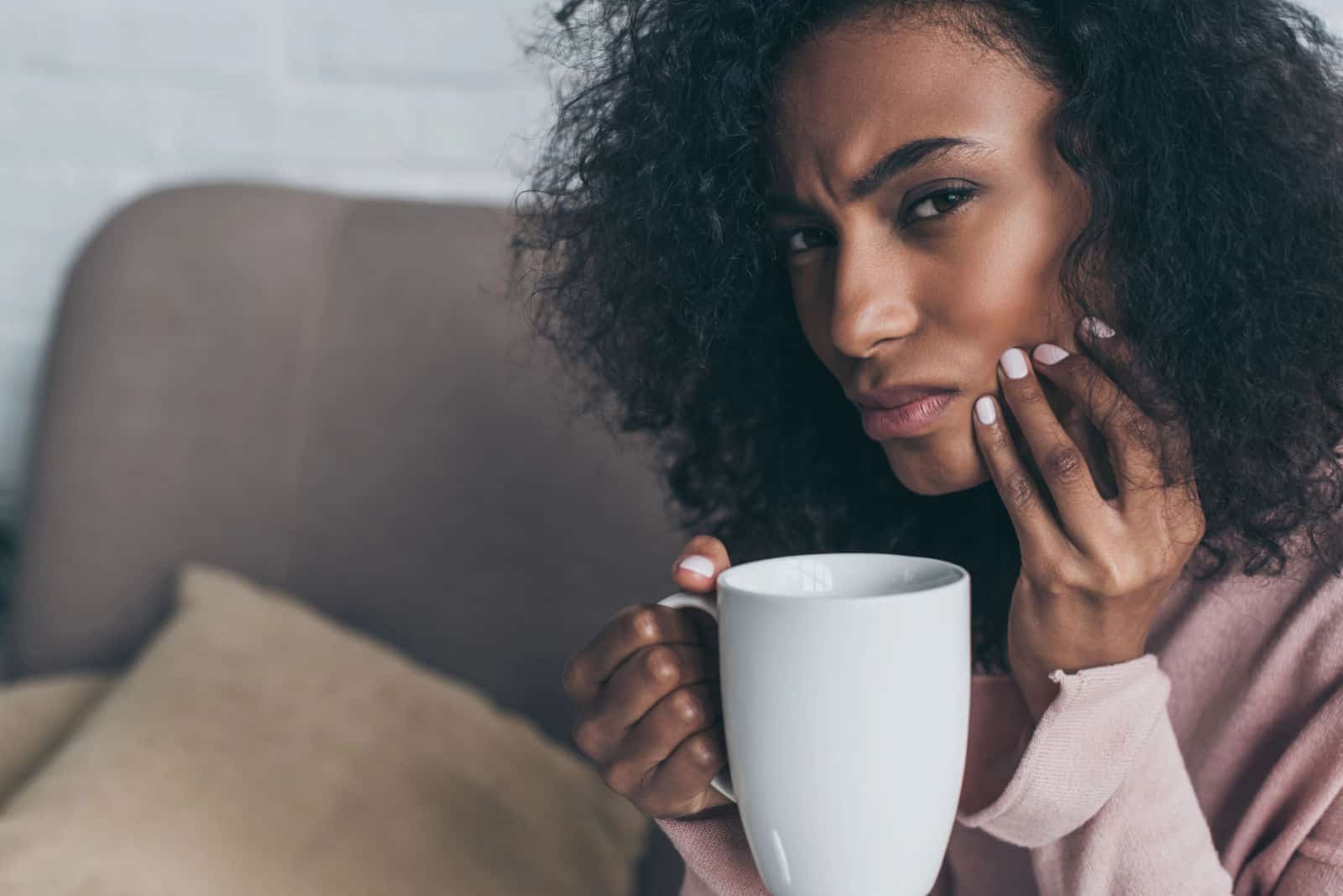 A young black woman holds her jaw because of TMJ pain while looking at the camera.