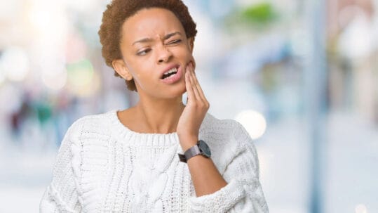 A young woman holds her jaw because of her TMJ pain in front of a blurred background.