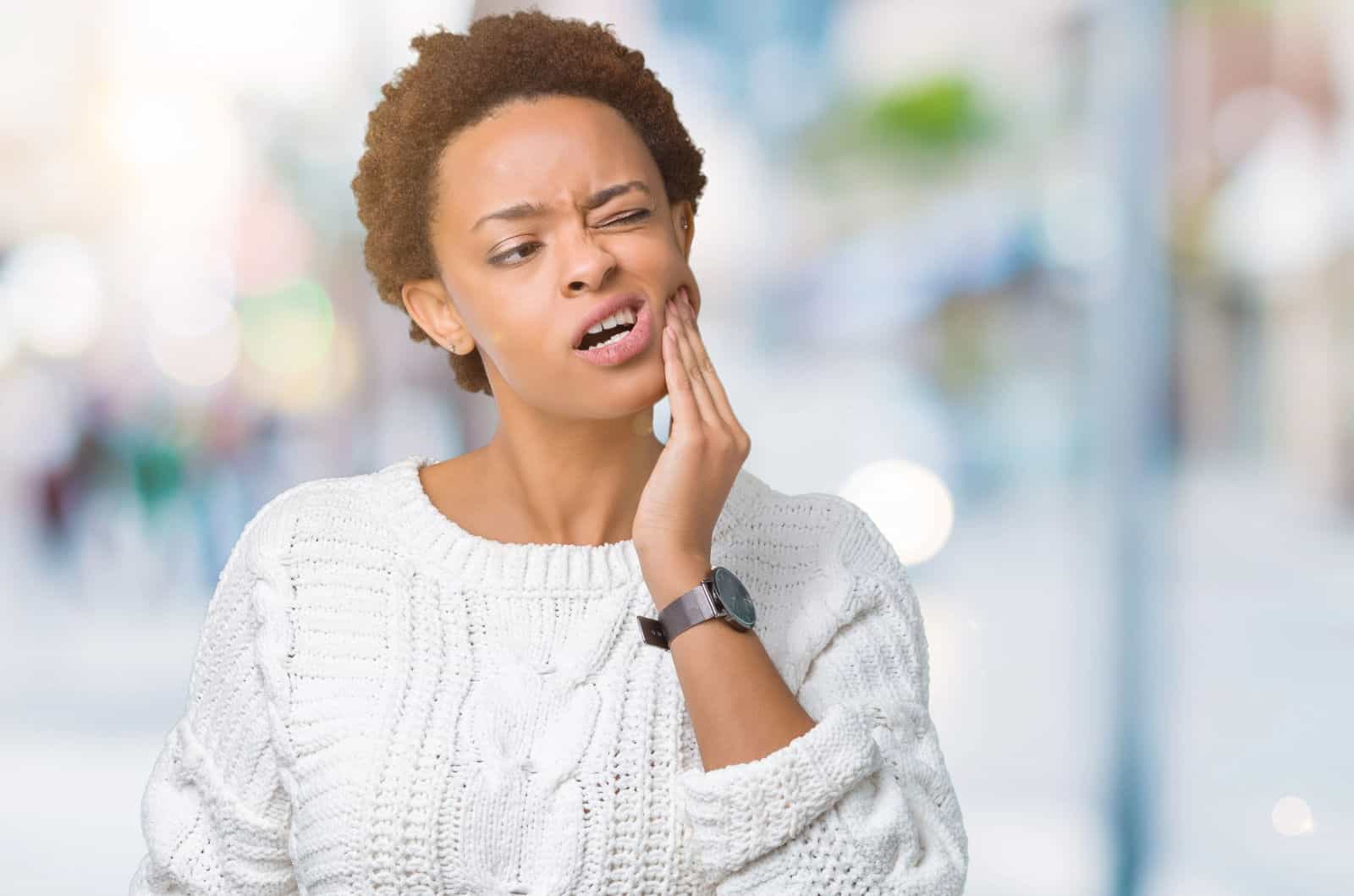 A young woman holds her jaw because of her TMJ pain in front of a blurred background.