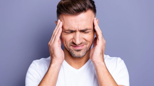 A man in a white v-neck holds his temples because his TMJ is causing a headache.