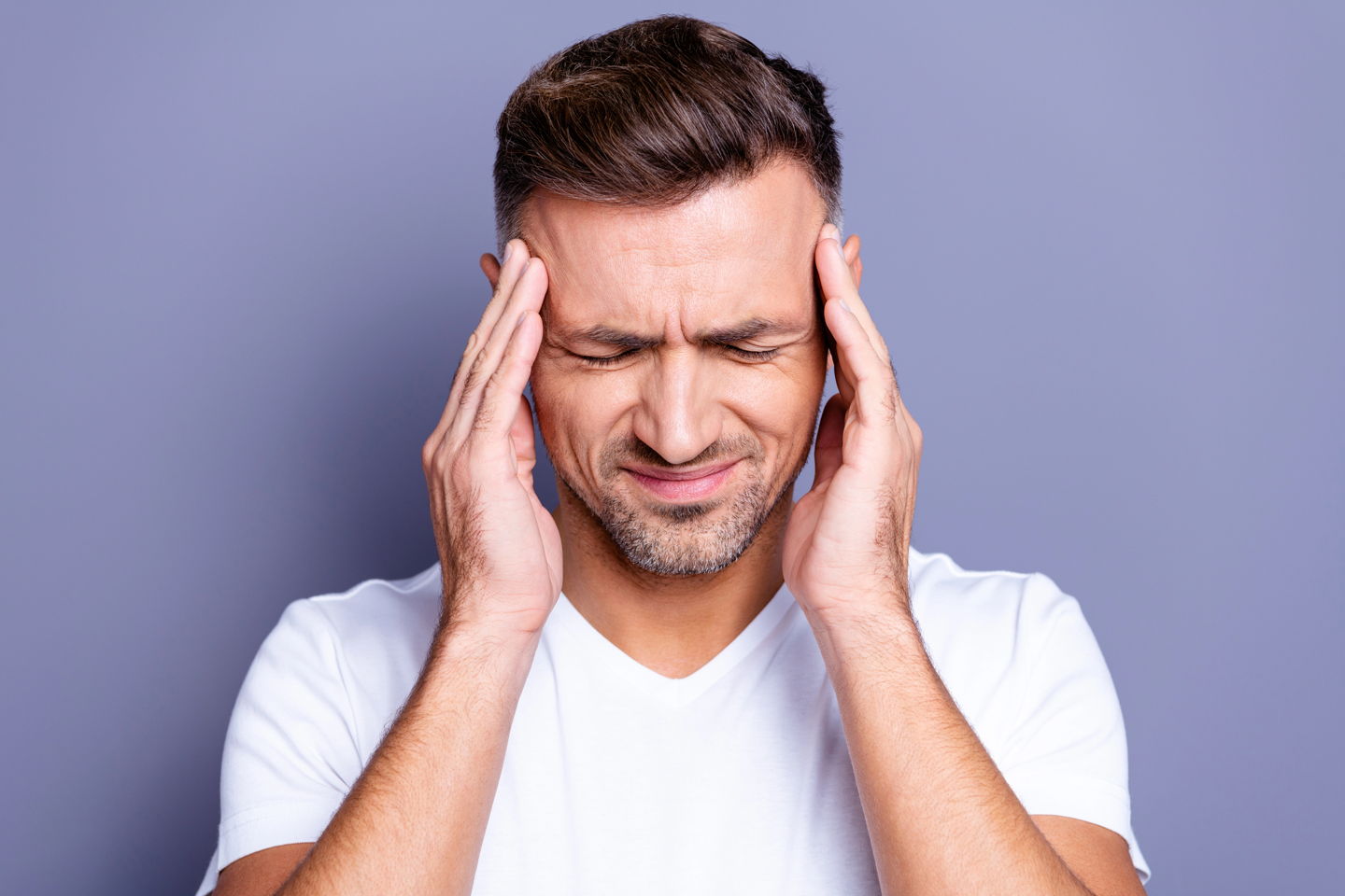 A man in a white v-neck holds his temples because his TMJ is causing a headache.